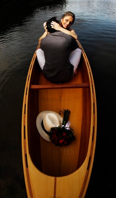 Vista de cima de um casal abraçado dentro de uma canoa de madeira, com o buquê e um chapéu ao lado.