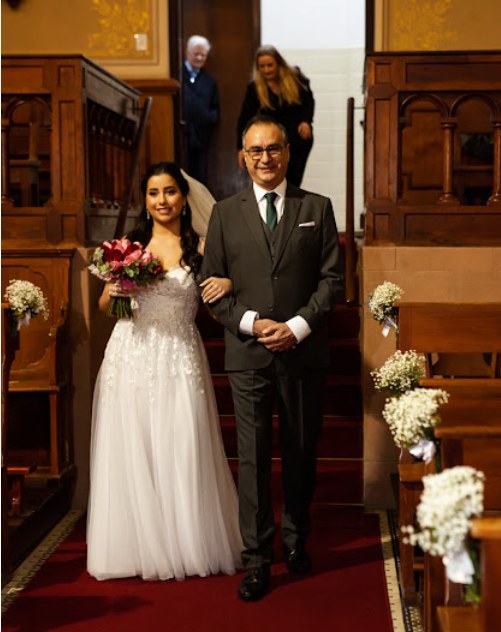 Noiva de braço dado com o pai descendo o corredor de uma igreja com tapete vermelho e bancos de madeira.