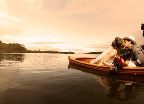 Casal de noivos se beijando dentro de uma canoa sobre a água espelhada, contra um pôr do sol dourado.